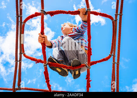 Blonde junge thront auf einem Web strickleiter Struktur in ein Kinderspielplatz für Spaß klettern. Stockfoto