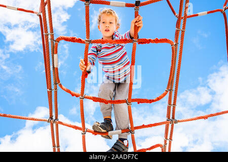 Blonde junge thront auf einem Web strickleiter Struktur in ein Kinderspielplatz für Spaß klettern. Stockfoto