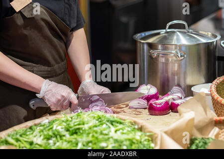 Cook's Hände in Zellophan Handschuhe schneiden rote Zwiebeln in dünne Scheiben. Kochen Gemüse Zutaten. Vor dem Hintergrund der Salatblätter und Pfannen Stockfoto