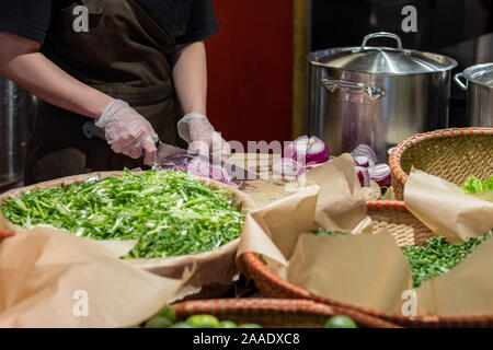 Cook's Hände in Zellophan Handschuhe schneiden rote Zwiebeln in dünne Scheiben. Kochen Gemüse Zutaten. Vor dem Hintergrund der Salatblätter und Pfannen Stockfoto