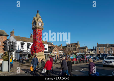 Menschen mit Fußgängerzone und Marktplatz Uhr dekoriert mit gestrickten Mohnblumen für Tag der Erinnerung Thirsk North Yorkshire England UK Vereinigtes König Stockfoto