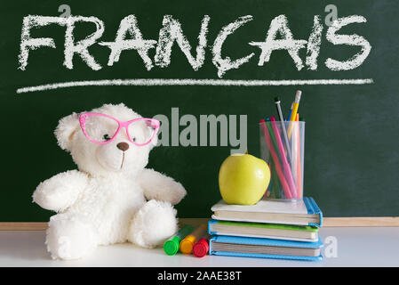 Teddy Bear mit Brille in der französischen Sprache im Klassenzimmer neben einen Apfel und einen Stapel Bücher und ein Wörterbuch gegen eine Tafel. Stockfoto