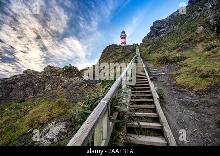 Blick auf Cape Palliser, rot-weiss gestreifte Leuchtturm mit steilen Treppen. Leuchtturm sitzt auf felsigen Grat mit üppiger Vegetation. Stockfoto