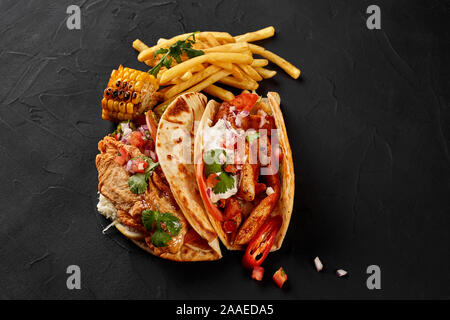 Chicken Fajitas mit gegrillten Zwiebeln und Paprika und mit Mehl Tortillas, Mais vom Grill und Pommes frites auf schwarzem Hintergrund. Ansicht von oben Stockfoto