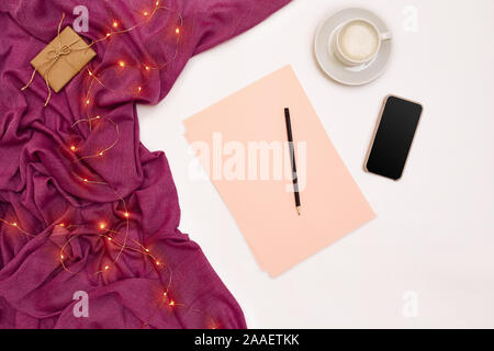 Eine Tasse Kaffee, schwarzer Smart, rosa Papier mit neuen Jahren Ziele. Schal und Weihnachtsbeleuchtung auf weißem Hintergrund. Stockfoto