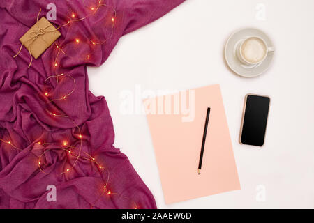 Eine Tasse Kaffee, schwarzer Smart, rosa Papier mit neuen Jahren Ziele. Schal und Weihnachtsbeleuchtung auf weißem Hintergrund. Stockfoto