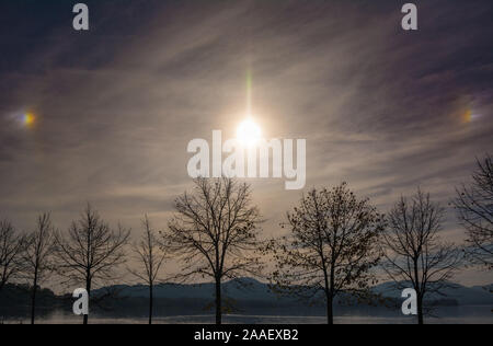 Sundogs plus eine Lichtsäule erscheinen in den Himmel über dem Lago Maggiore, Italien in kalten Bedingungen im frühen Winter. Stockfoto