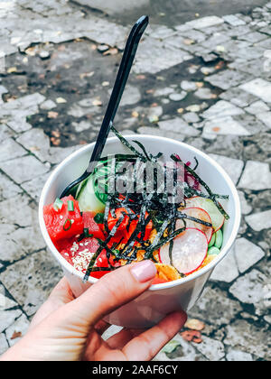 Poke Schüssel mit Thunfisch in das verfügbare Papier container. Frau hand mit traditionellen Hawaiianischen fast food auf einer Straße der Stadt. Mobile Fotografie Stockfoto