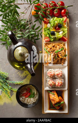 Komplettes Mittagessen. Griechischer Salat, Glasnudeln, Brötchen, Lachsfilet, Nudelsuppe und Teekanne mit Tee auf einem grauen Hintergrund. Die Zusammensetzung ist eingerichtet w Stockfoto