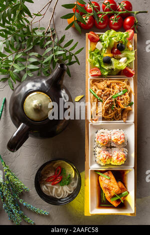 Komplettes Mittagessen. Griechischer Salat, Glasnudeln, Brötchen, Lachsfilet, Nudelsuppe und Teekanne mit Tee auf einem grauen Hintergrund. Die Zusammensetzung ist eingerichtet w Stockfoto