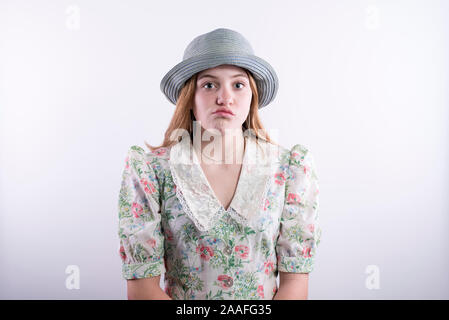 Kaukasische teenage Mädchen mit einem Vintage geblümten Kleid und blauen Hut mit gelangweilten Blick auf ihrem Gesicht vor einem weißen Hintergrund Stockfoto