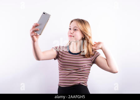 Kaukasische teenage Mädchen mit einem bunten, horizontal gestreifte T-Shirt und lächelnd, während sie einen selfie mit einem Smartphone vor einem weißen Hintergrund Stockfoto