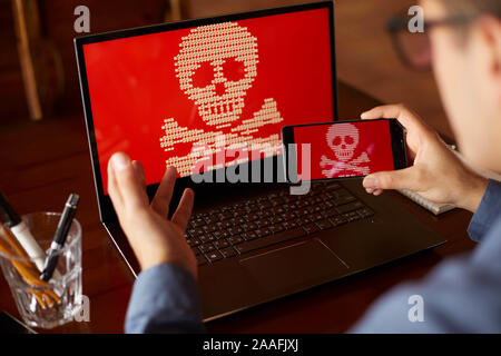 Mann sitzt in der Nähe von Laptop mit Telefon gesperrt und von ransomware Spyware um Geld bittet verschlüsselt. Laptop und Smartphone von Virus infiziert. Gruselig roten Schädel Stockfoto