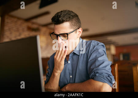 Am frühen Morgen im Büro. Schläfrig, müde aussehenden hipster Freelancer in Gläsern ist Gähnen an seinem Arbeitsplatz in vor dem Bildschirm auf dem Schreibtisch Stockfoto