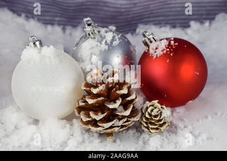 Drei Weihnachtsverzierungen und ein paar Kiefernzapfen Rest auf dieser winterlichen Hintergrund. Vereinzelt Schnee ziert die Ornamente, Kiefernzapfen und Hintergrund. Stockfoto