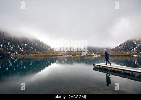 Einsamer Mann mit Rucksack und Kamera zu Fuß den Pier am nebligen See mit Reflexion an der Herbst in Kasachstan und Zentralasien Stockfoto