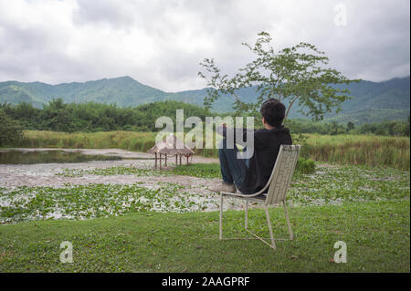 Mann, Jacke sitzt auf einem Stuhl im Garten mit Sumpf und Berg Stockfoto