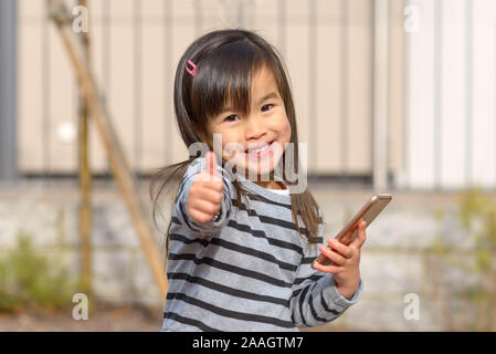 Cute glückliche junge Mädchen mit Grübchen lächelnd in die Kamera, als Sie einen Daumen nach oben Geste mit der Hand gibt, während Sie ein Handy im Freien in sunsh Stockfoto