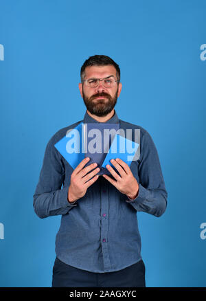 Professor mit ernsten Gesicht. Mann mit Bart und Bücher. Lehrer trägt eine Brille und Notebooks. Wissen und die unterrichtsplanung Konzept. Lehrbücher in blauer Farbe in der Kerle Hände auf blauem Hintergrund Stockfoto