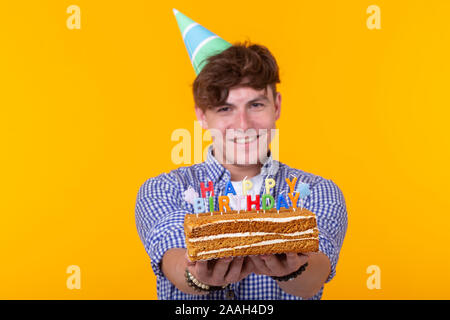 Positive junge Mann hält ein Happy birthday cake und zwei brennende bengalische Lichter auf gelbem Hintergrund posiert. Werbeflächen Stockfoto