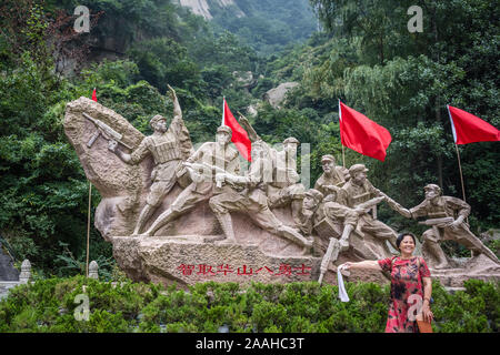 Huashan, China - August 2019: Chinesische Frau vor historischen Skulptur Darstellung kommunistischen Soldaten mit Waffen posiert anbd rote Fahnen auf der Stockfoto