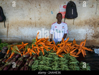 Ein Mann mit einem Liverpool Football Shirt steht hinter seinem Marktstand verkaufen, Möhren, Bohnen und Auberginen in Mzuzu Markt, Malawi. Stockfoto