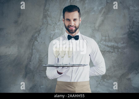 Foto fröhliche, positive toothy strahlende Kellner mit Tablett mit Gläsern Sekt trinken über graue Betonwand Farbe Hintergrund isoliert Stockfoto