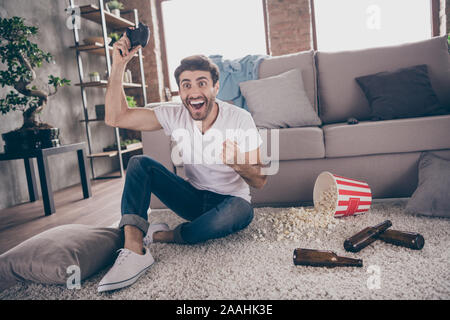 Foto von gemischten Rennen arabischen Kerl sitzen Teppich in der Nähe von Sofa halten Joystick spielen video Spiel aufgeregt gewinnen die Fäuste leeren Bierflaschen Popcorn auf dem Boden Stockfoto