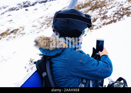 Sport Mädchen Snowboarder in Helm und Maske nimmt Bilder mit Smartphone, sitzen auf Kabel weg. Konnektivität Mobilität lifestyle Technologie. Reisen L Stockfoto