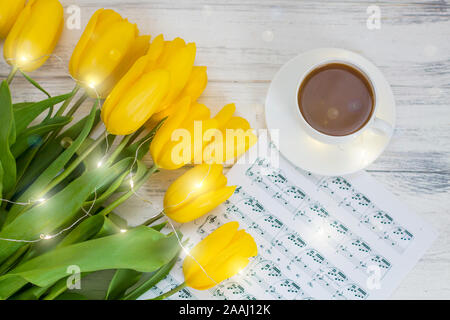 Gelbe Tulpen mit Girlanden, eine Tasse Kaffee, Cappuccino und Notizen auf einem weißen Holz- Hintergrund Stockfoto