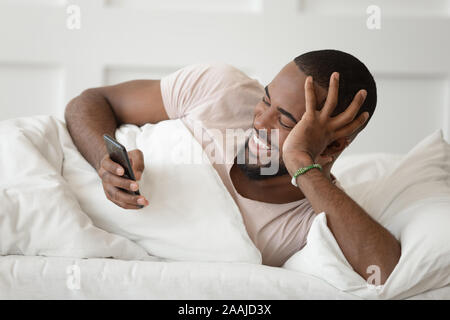 Happy afrikanischer amerikanischer Mann im Bett liegend mit Handy Stockfoto