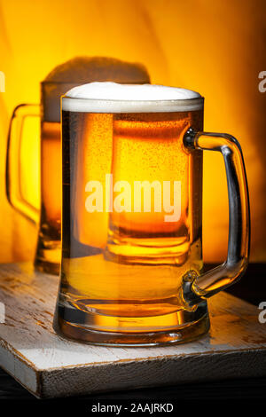 Zwei helle Bier Becher mit weißer Schaum auf dunklen Holztisch und gelbem Hintergrund Stockfoto