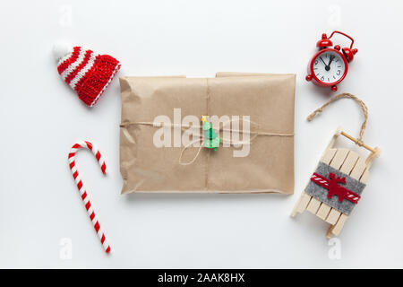 Weihnachten Geschenk verpackt in braun Handwerk Papier, mit der geissel gebunden, mit Zuckerrohr Süßigkeiten, kleine analoge Uhr, Schlitten, Hut auf weißem Hintergrund. Minimalistischer Stil. Top Stockfoto