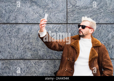 Exzentrische bärtiger Mann mit Platinum blonde Haare und Sonnenbrille nimmt Bilder mit seinem Smartphone , Hipster posieren für selfie Stockfoto
