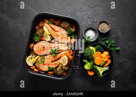 Lachs. Gebackenen Lachs Fisch Steaks mit Gemüse, Brokkoli, Karotten und Zwiebeln auf schwarzem Hintergrund, Ansicht von oben Stockfoto