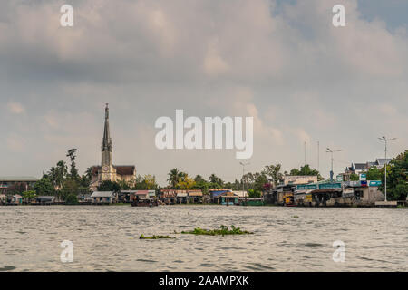 Cai, Mekong Delta, Vietnam - März 13, 2019: Entlang der Kinh 28 Kanal. Landschaft mit Katholische Kirche hinter Reihe von Einzelhandelsunternehmen und Grünen fo Stockfoto