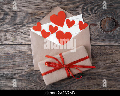 Karte und rote Herzen in offenen Umschlag aus braunem Kraftpapier. Geschenkbox mit roter Schleife auf hölzernen gealterten vintage Hintergrund. Grüße mit Valentinstag da Stockfoto