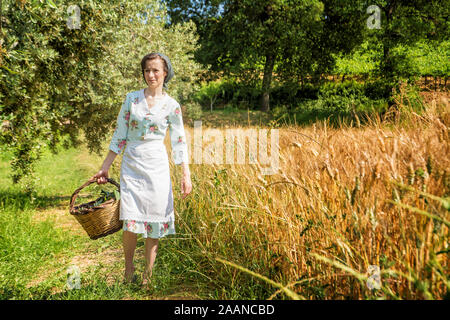 Frau in 40 s Kleider Spaziergänge in der italienischen Landschaft, neben einem Weizenfeld, ein Korb mit Kirschen Stockfoto