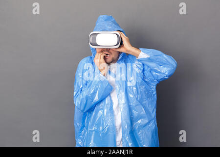 Unterhaltung. Junger Mann im Regenmantel und Virtual Reality headset Studio stehen auf grau Video isoliert Angst Stockfoto