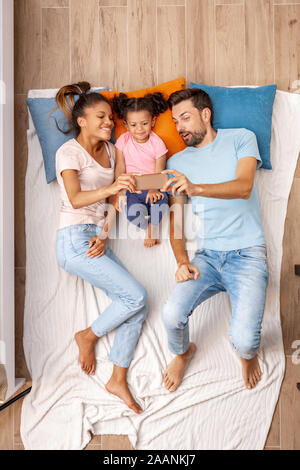 Mischlinge Familie. Mutter, Vater und Tochter auf dem Bett liegend unter selfie am Telefon lächeln, freundliche Ansicht von oben Stockfoto