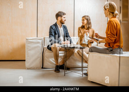 Junges Paar im Gespräch mit einem Sales Manager oder Immobilienmakler, sitzend auf der bequemen Couch im Büro Stockfoto