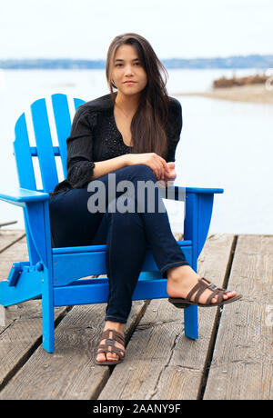 Biracial Thai Kaukasischen jugendlich Mädchen in blau Adirondack Stuhl sitzen auf hölzernen Pier am See im Freien Stockfoto