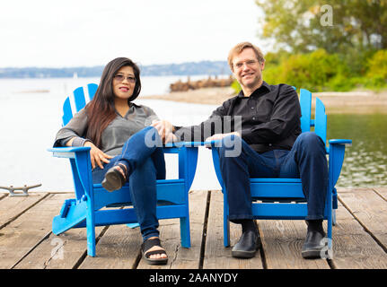 Gemischtrassiges Paar, kaukasischer Mann und die asiatische Frau, sitzen auf den Blauen andirondack Stühle auf hölzernen Pier am See im Freien im Sommer Stockfoto