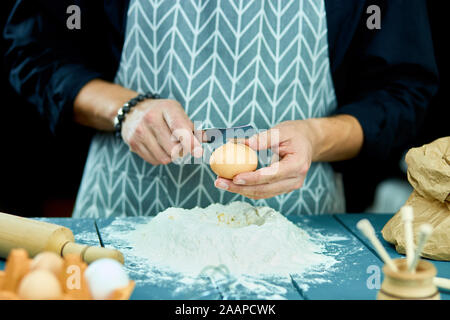 Aufschlagen der Eier in das Mehl. Nahaufnahme der männlichen Hände brechen rohes Ei über Mehl. Baker kneten den Teig für Kuchen. Der Prozess der Aufschlagen der Eier. Stockfoto