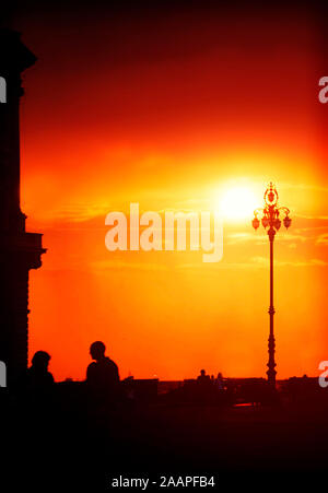 Roter Himmel bei Sonnenuntergang über dem Meer promenade und unita' d'italia Square in Triest, Italien mit Silhouetten von einem Gebäude und Menschen im Licht Stockfoto