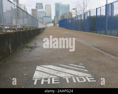 "Die Flut" im Schriftzug auf den Gehweg für Fußgänger und Radfahrer in der oberen Flussufer der Greenwich Peninsula, mit Canary Wharf in der Ferne. Stockfoto