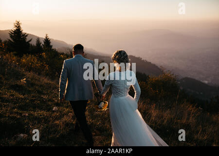 Wunderschönes Paar, das einen romantischen Moment an ihrem Unkraut, in den Bergen bei Sonnenuntergang, hat. Miteinander laufen. Die Frau ist in einem weißen Hochzeitskleid. Stockfoto