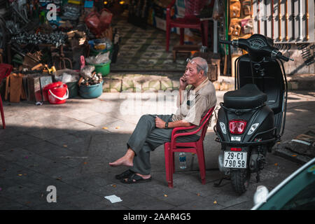 Hanoi, Vietnam - 12. Oktober 2019: Eine alte ältere Menschen asiatischer Mann sitzt auf einem Stuhl aus Kunststoff neben einem Moped auf dem Telefon Stockfoto