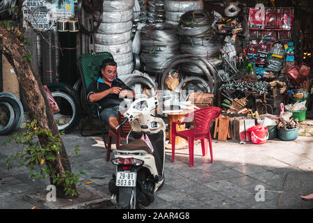 Hanoi, Vietnam - 12. Oktober 2019: Ein asiatischer Mann sitzt auf einem Stuhl neben einem Moped vermeiden der heißen Hitze des Tages texting stürzte Stockfoto