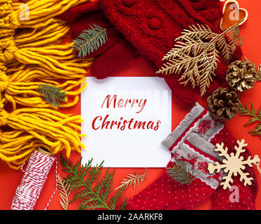 Frohe Weihnachten Karte. Winter Socken, gestrickte Schal und Handschuhe. Zweige. Roten Hintergrund. Flach, Ansicht von oben. Saisonale warme Kleidung. Stockfoto